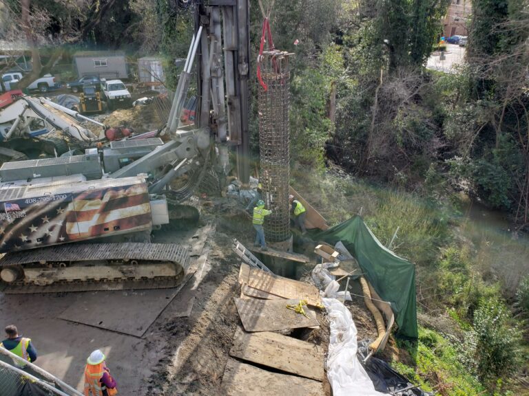 A construction crew using a large drilling rig crane to lower a massive cylindrical steel rebar cage into a deep foundation shaft for a caisson installation.